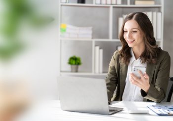 Junge Frau im Büro am Laptop mit Handy in der Hand
