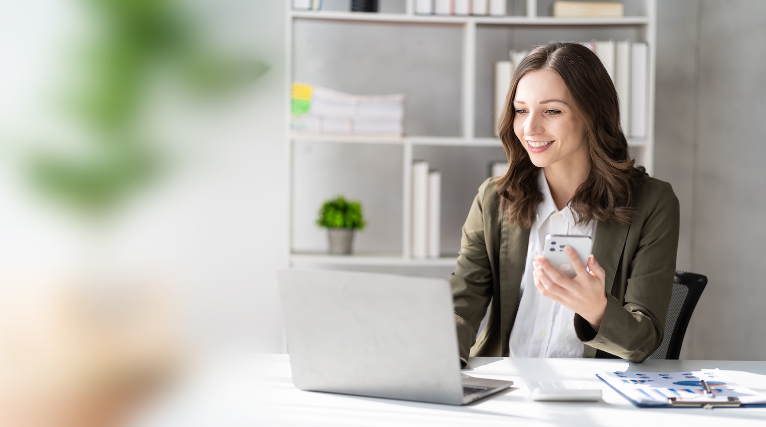 Junge Frau im Büro am Laptop mit Handy in der Hand